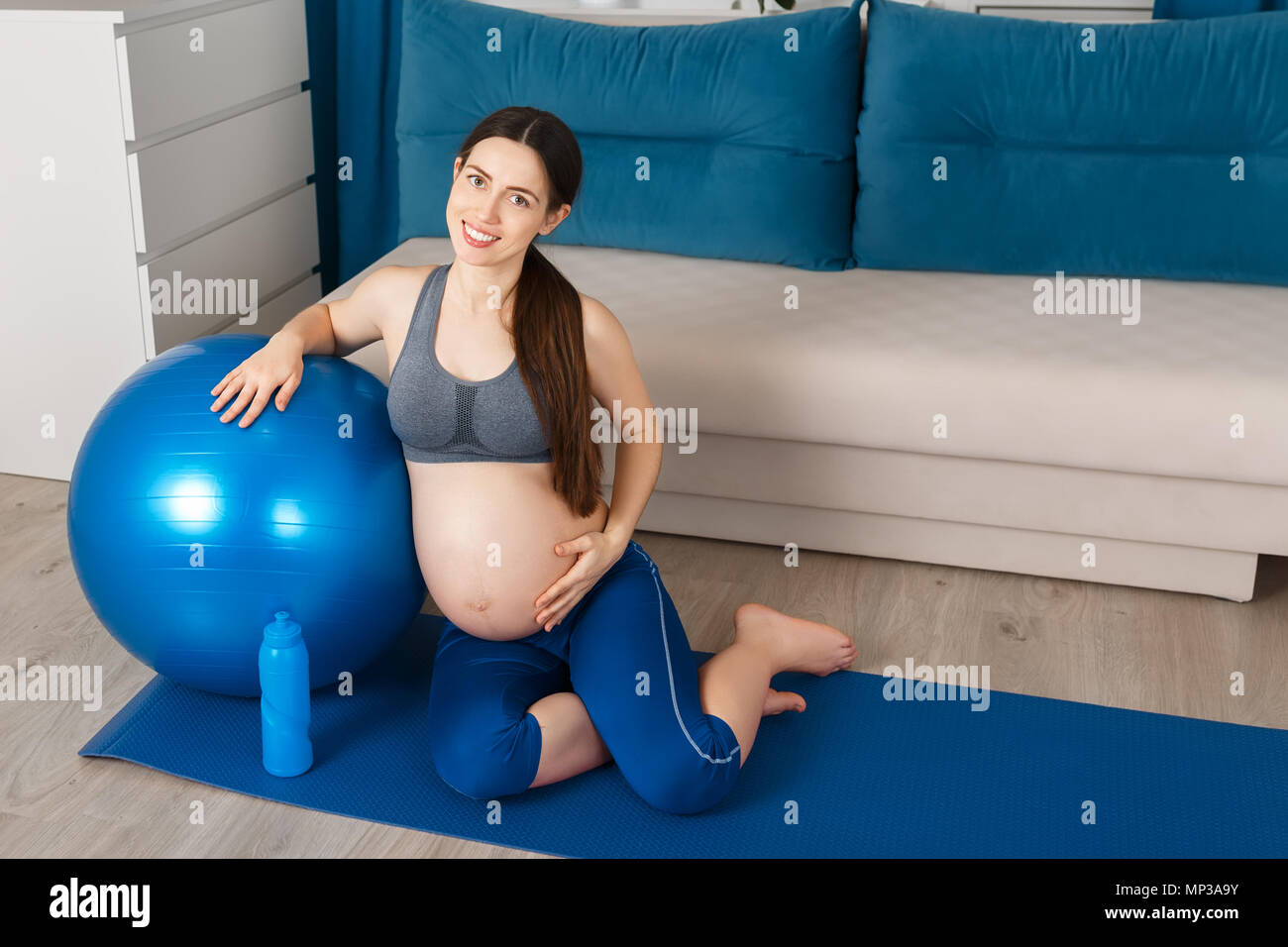 Glücklich schwanger Frau mit blauen Fit Ball und einer Flasche Wasser sitzen auf Yoga Matte. Die werdende Mutter ihr Bauch berühren nach Gymnastik zu Hause Stockfoto