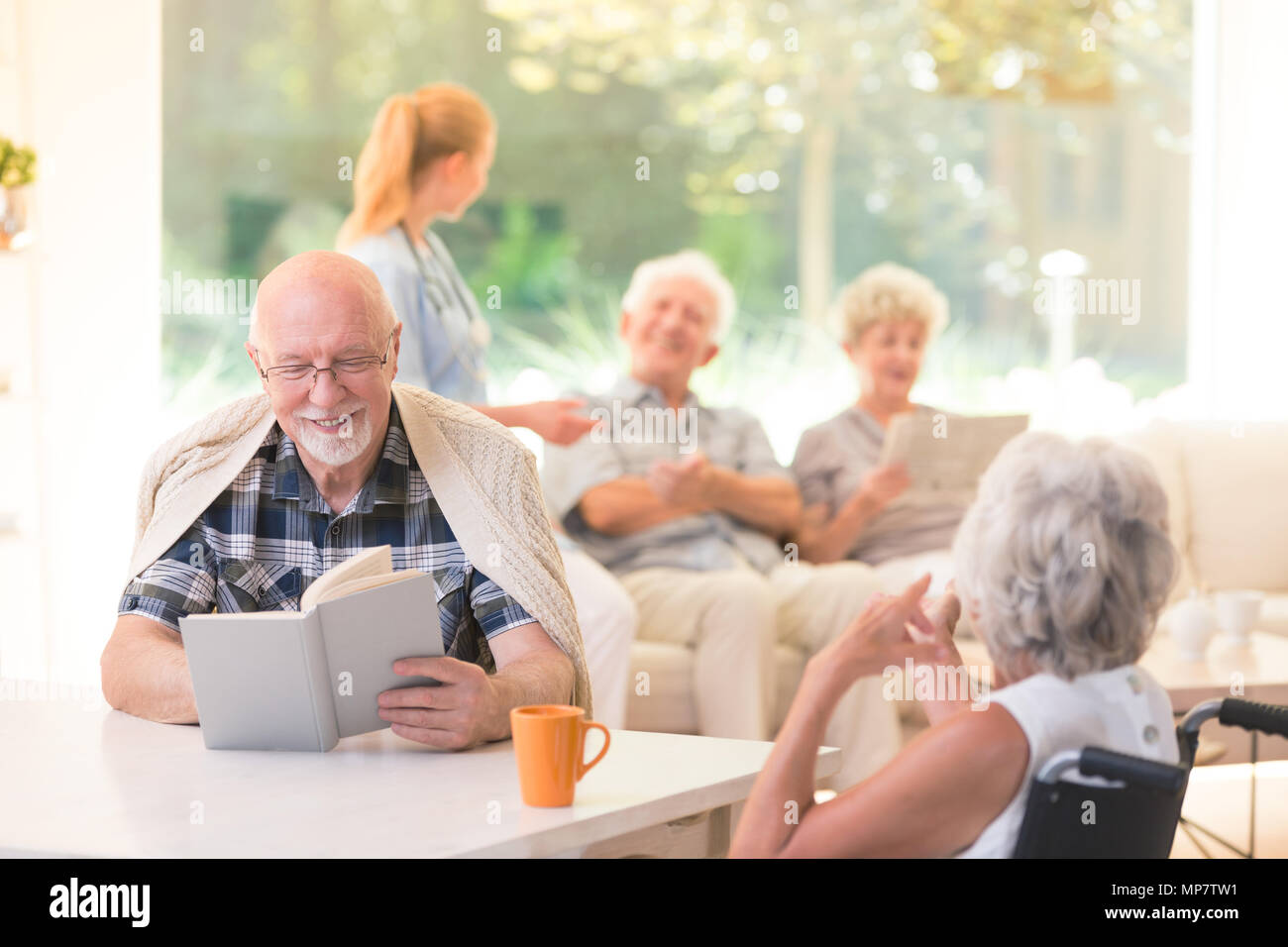 Älterer Mann ein Buch lesen und lächelnd, während sie mit einem Freund an einem Tisch in der Krankenpflege Haus Stockfoto