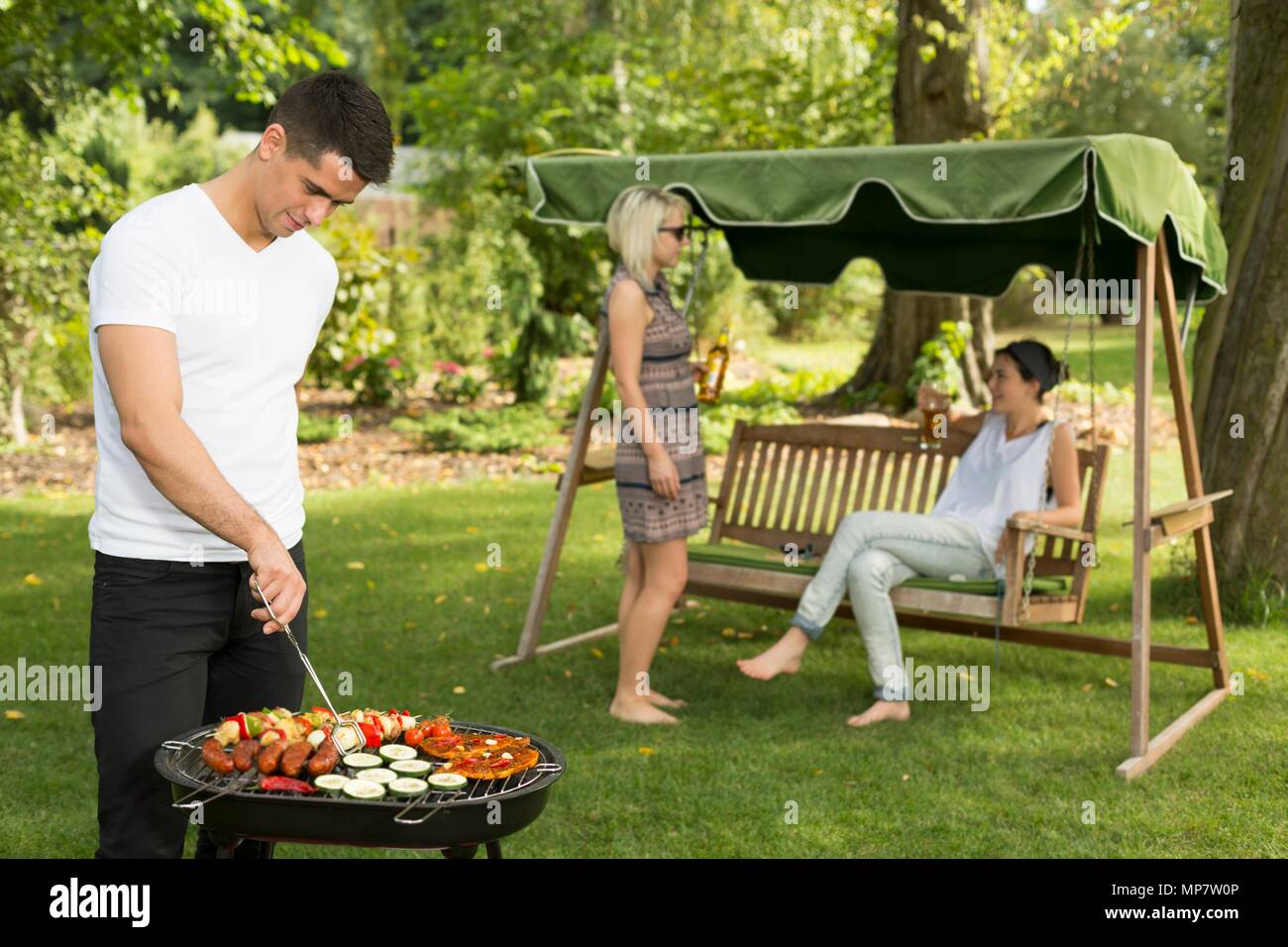Aufnahme eines jungen Mannes grillen Essen und seine Freunde sprechen im Hintergrund Stockfoto