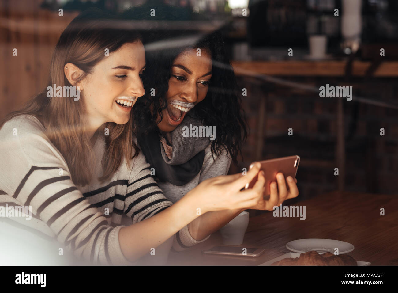 Zwei Frauen, die eine selfie lachend und an ihre Milch Schnurrbärte im Coffee Shop. Freunde sitzen in einem Café mit Kaffee und Snacks auf der Tabelle t Stockfoto