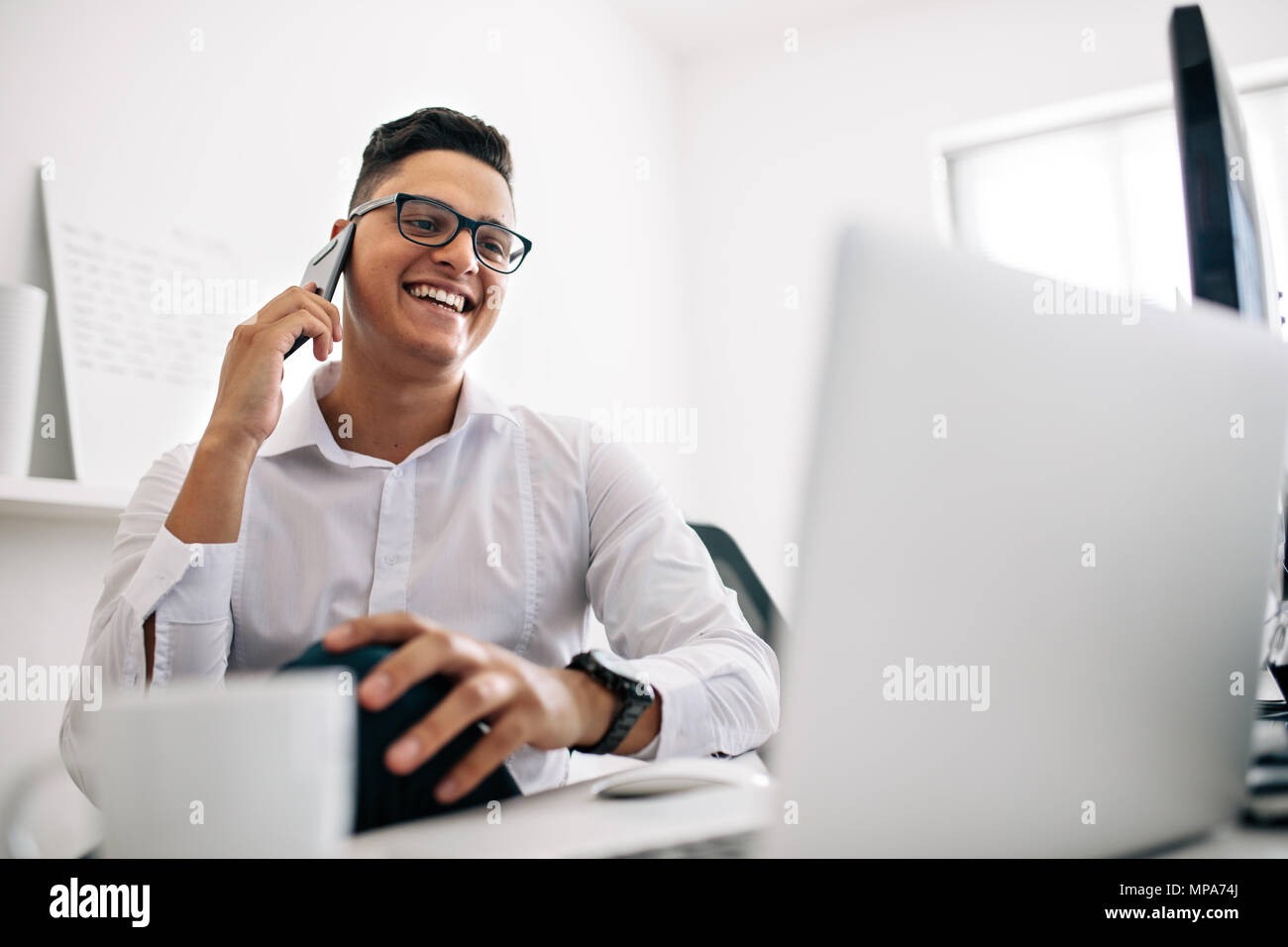Lächelnd Software Entwickler sprechen über Handy in seinem Büro Schreibtisch mit einem Laptop und der Kaffeetasse in den vorderen Sitzen. Mann mit Brille arbeiten Stockfoto