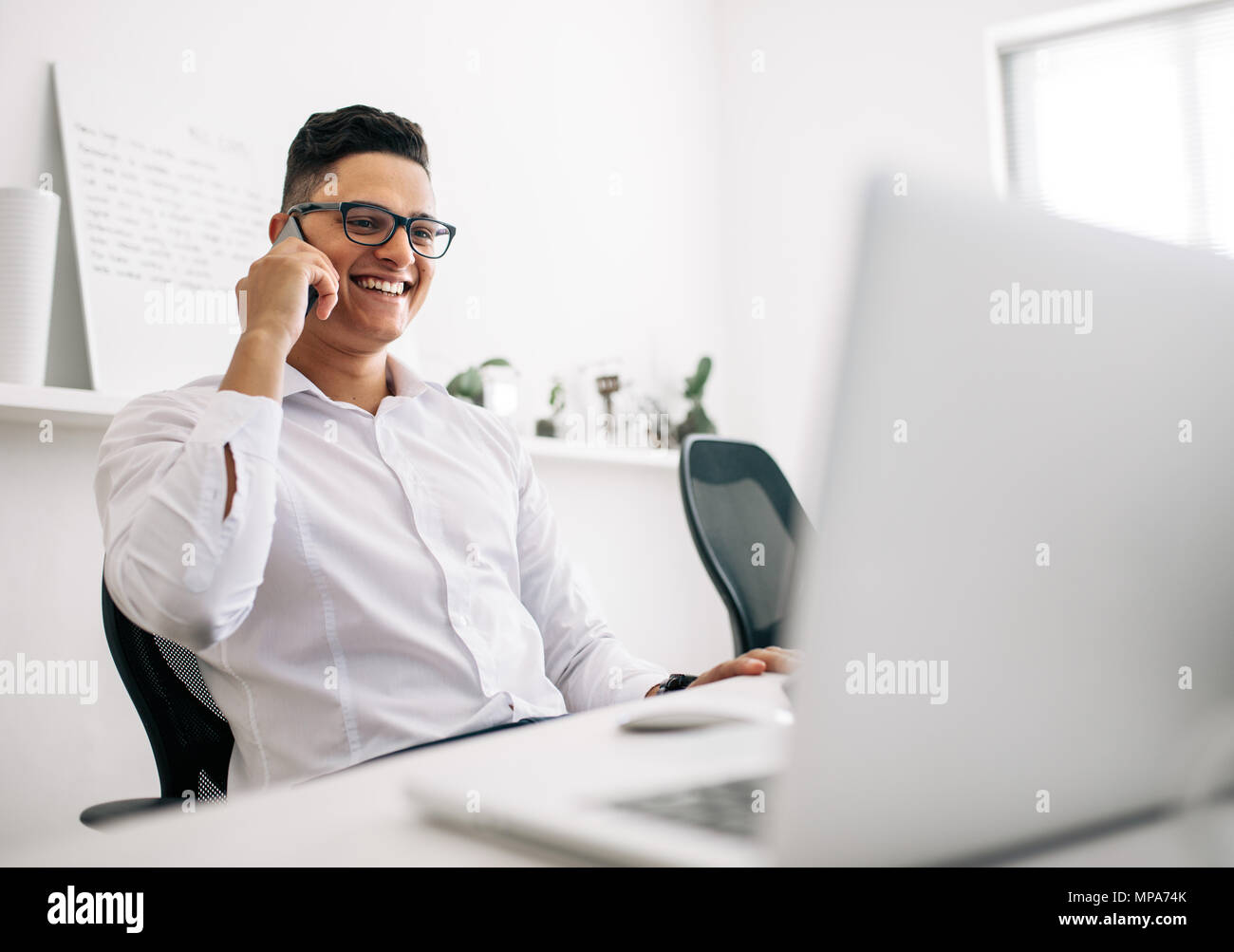 Lächelnd Software Entwickler sprechen über Handy an seinem Schreibtisch mit einem Laptop in der vorderen Sitzen. Mann mit Brille arbeiten am Laptop compu Stockfoto