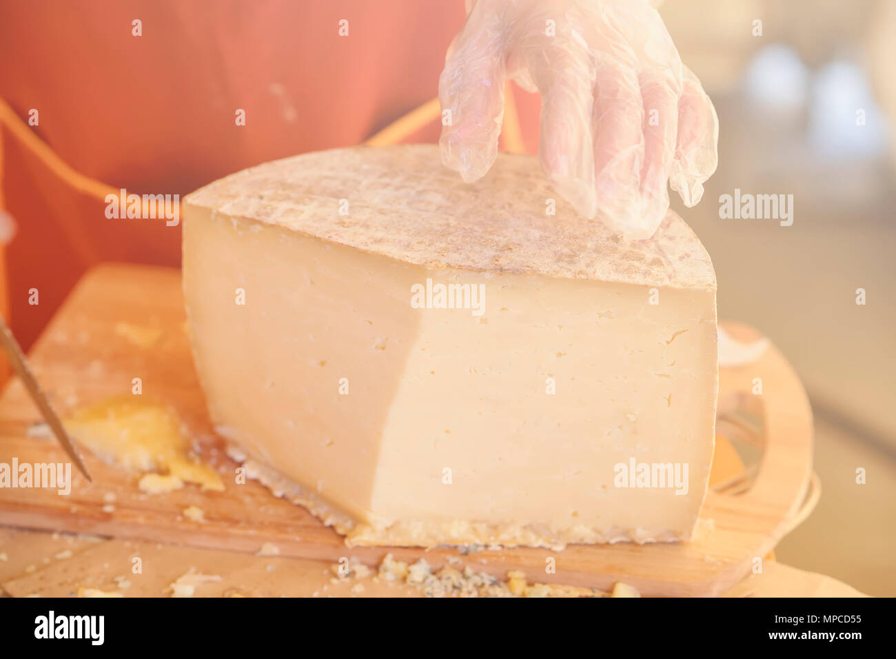 Die Hände des Verkäufers in Handschuhen und die Hälfte von einem großen Kopf von Käse close-up, Holz- Board. Selektive konzentrieren. Gastronomische Milchprodukte, realen Szene, essen Markt Stockfoto