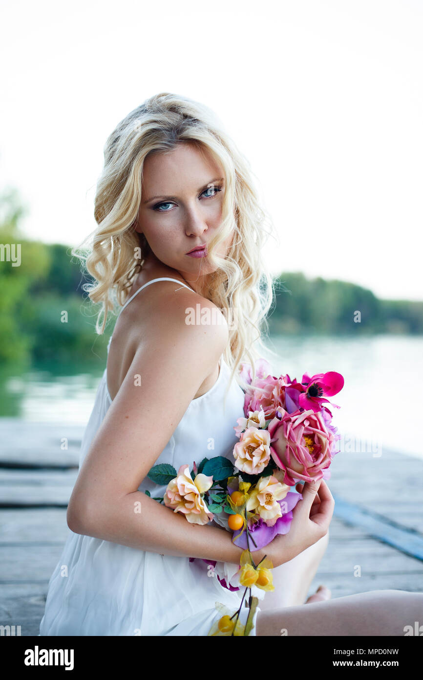 Junge schöne blonde Frauen mit blauen Augen und welliges Haar lang mit der Natur Hintergrund. Slawische Frau mit romantischen weißen Kleid und mit Blumen Stockfoto