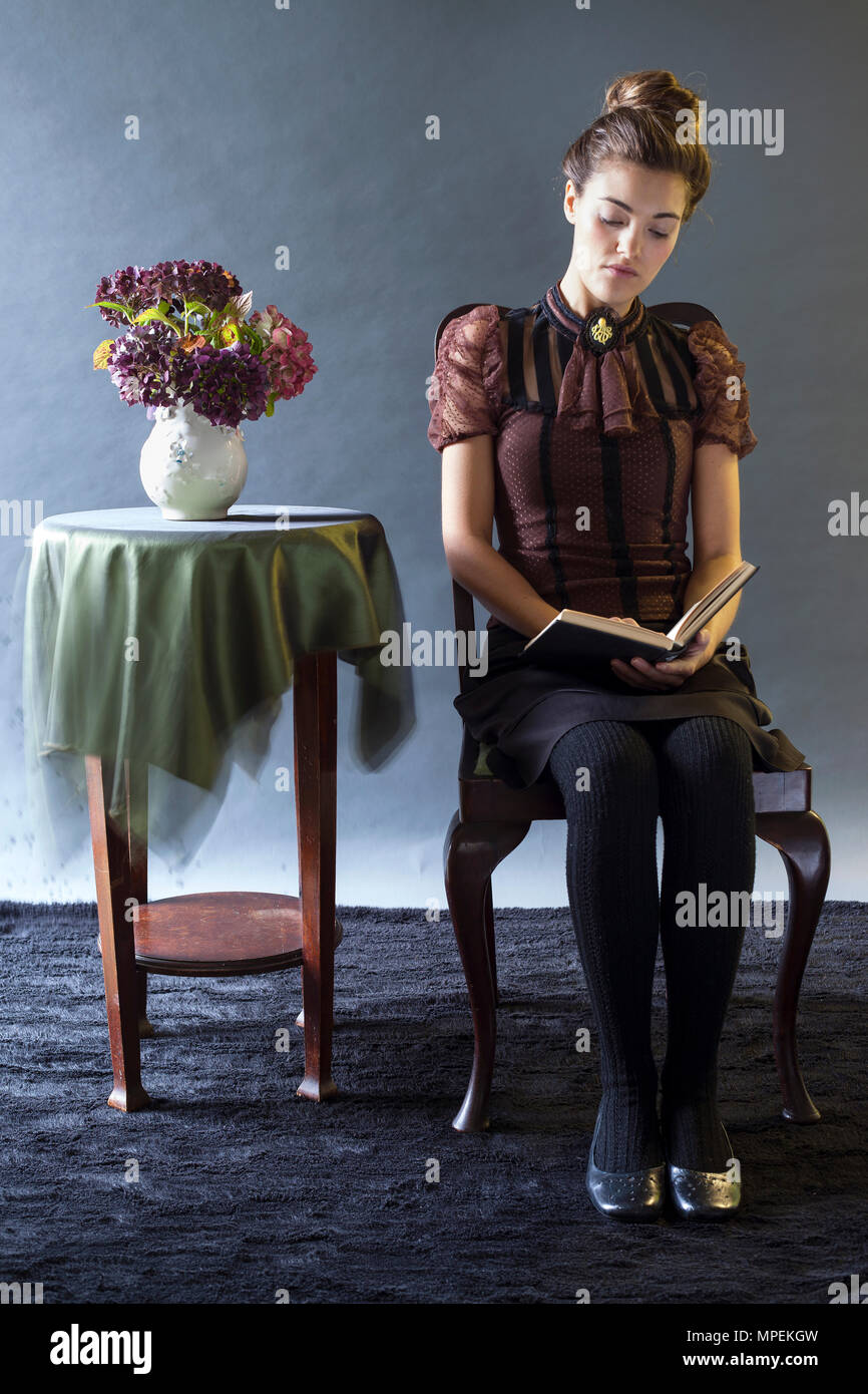 Junge Frau in Vintage-Mode Bluse sitzt neben dem Tisch mit Blumen in Vase lesen, Modell Release zur Verfügung Stockfoto