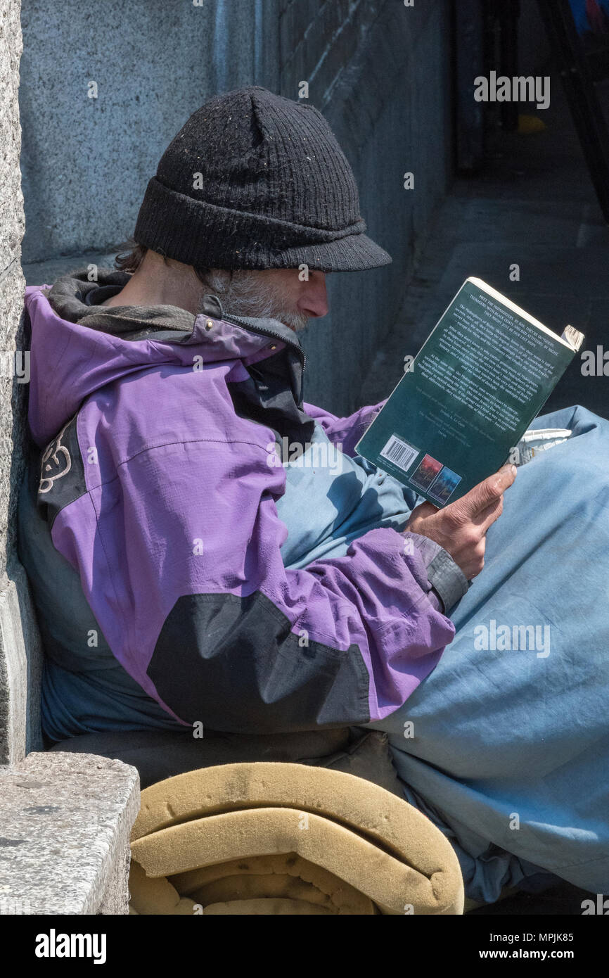 Ein obdachloser Mann sitzen auf dem Boden trägt einen schwarzen Hut und ein Buch lesen im Zentrum der Hauptstadt von London. obdachlosigkeit in der Hauptstadt. Stockfoto