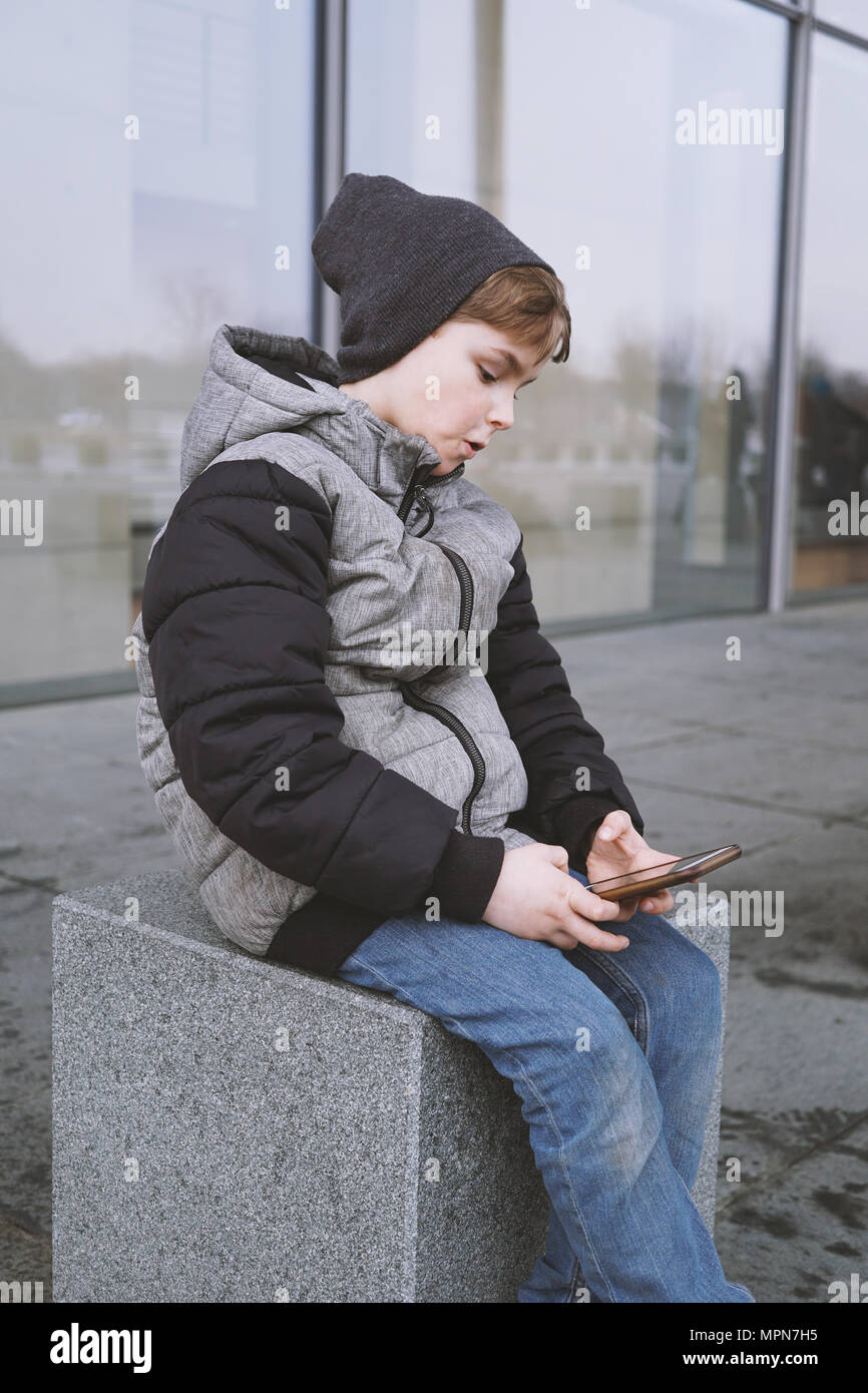 7 Jahre alten Junge spielt mit Smartphone, Draußen sitzen im Winter Jacke und Mütze stricken Stockfoto