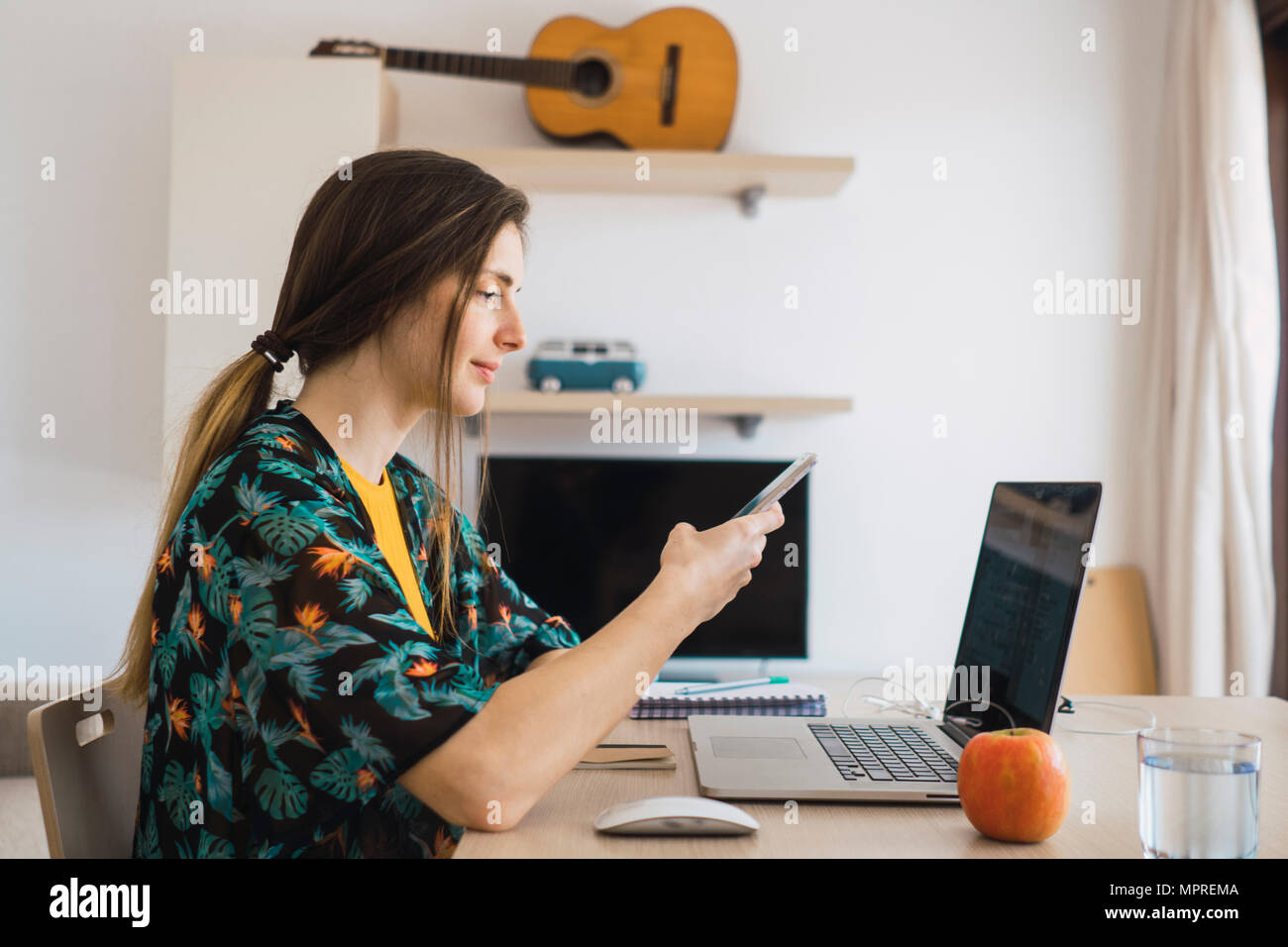 Junge Frau am Tisch zu Hause mit Handy und Laptop Stockfoto