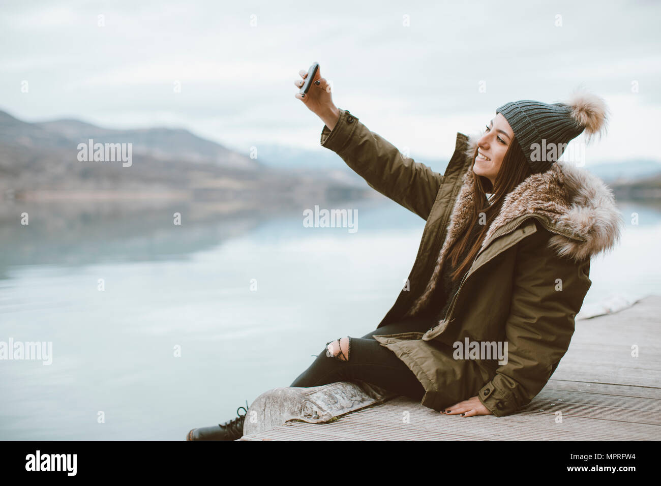 Lächelnde junge Frau sitzt auf der Jetty unter selfie mit Smartphone Stockfoto