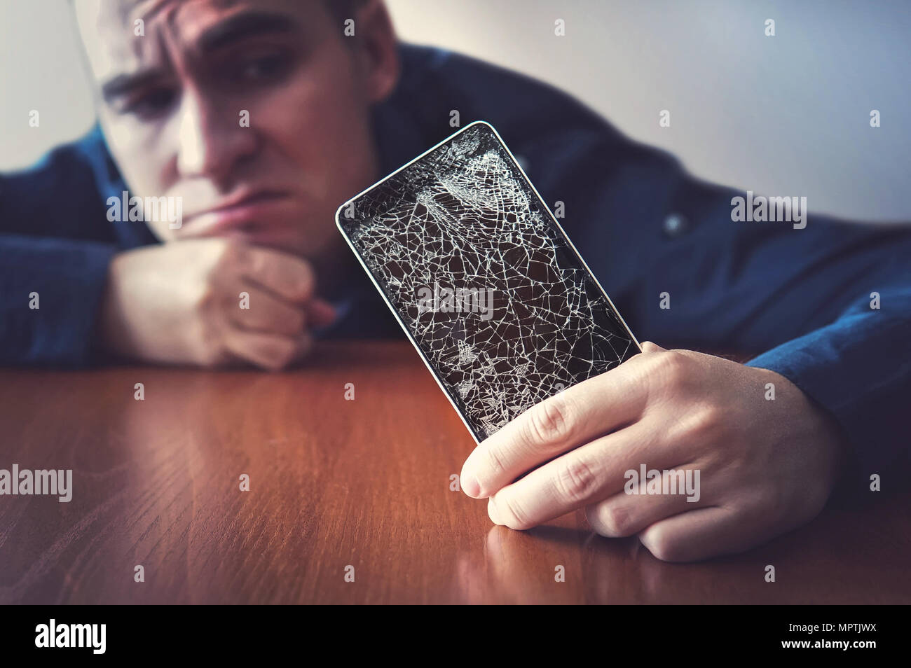 Hände halten ein Mobiltelefon mit ein zerbrochenes Display über die hölzerne Oberfläche. Ein Mann schaut auf seinem gebrochenen Telefon liegen auf einem Holztisch mit einem traurigen Blick. gu Stockfoto