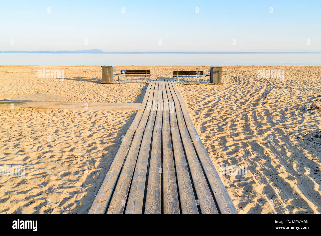 Holzsteg an einem Strand, mit zwei leeren Bänken und Mülleimer an den Seiten. Windstille Meer unter dem Horizont, an einem sonnigen Morgen, bevor die Badenden ar Stockfoto