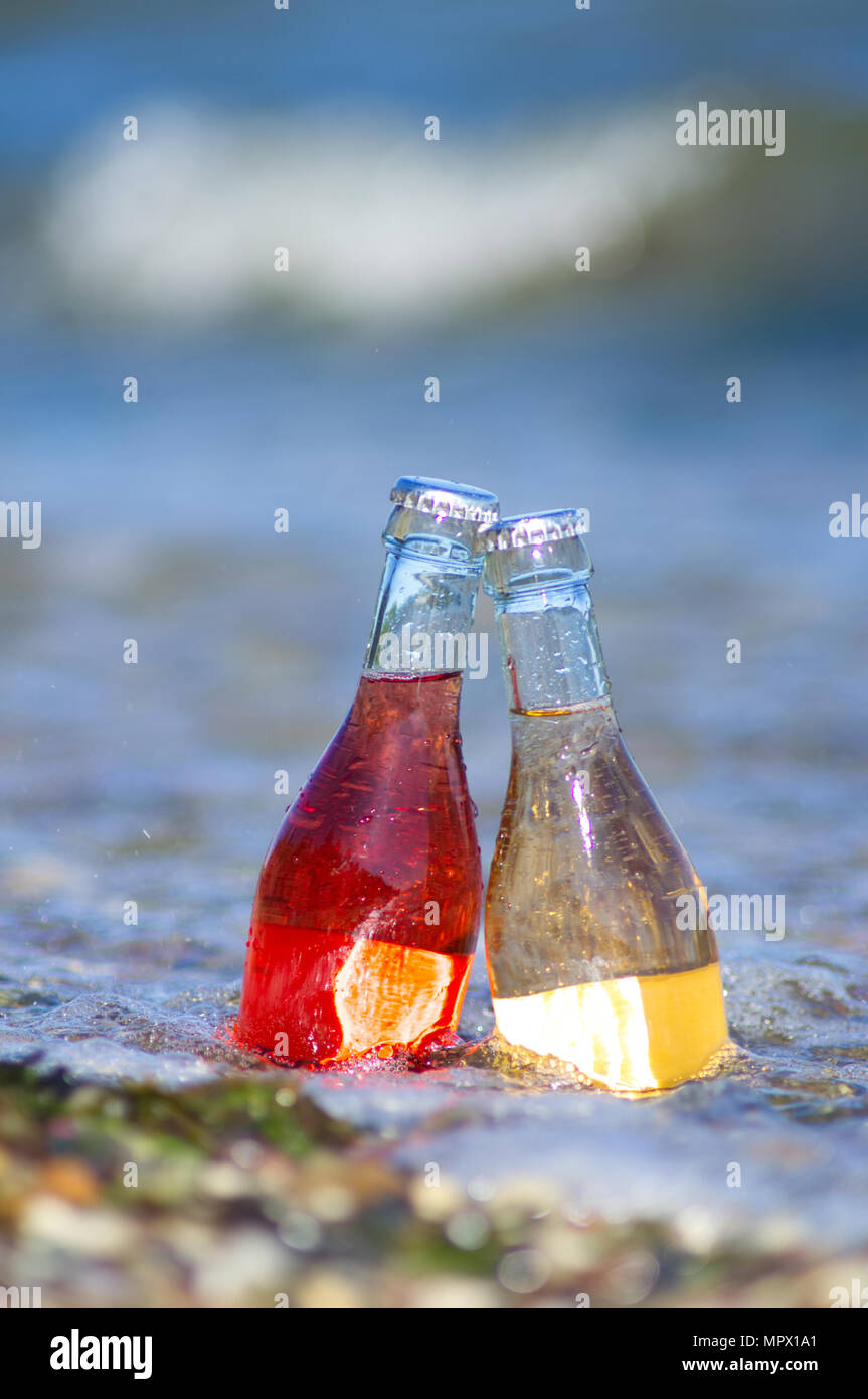 Zwei Flaschen stehen im Wasser am Strand im Sommer. Rote und gelbe Getränke. Stockfoto