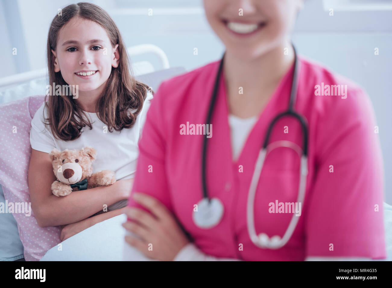 Lächelnd krankes Mädchen mit Plüsch Spielzeug. Arzt in rosa Uniform im Vordergrund, unscharf Stockfoto