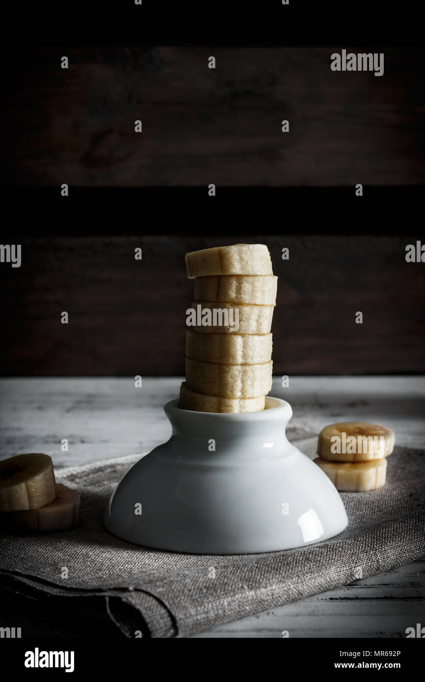 Köstliches Dessert frischen in Scheiben geschnittenen Banane stehen auf einer weißen Schüssel mit Segmente um auf einem Sack und in der Holz- Hintergrund. Vertikale Bild der dunklen Stimmung Stockfoto