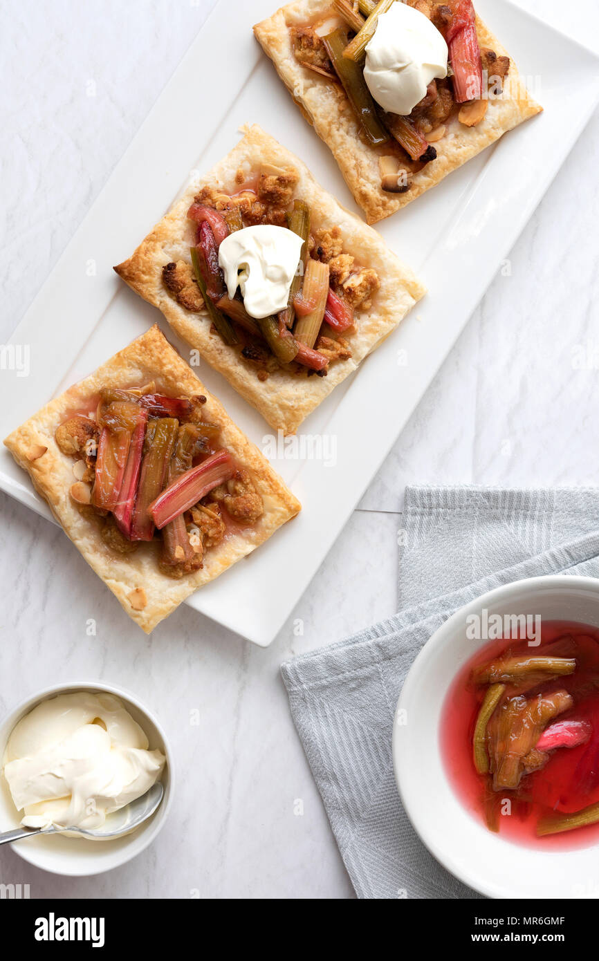 Drei kleine Quadrat Rhabarber und Mandel Kuchen mit einer Schüssel mit Sahne und Schüssel gekochten Rhabarber. Stockfoto