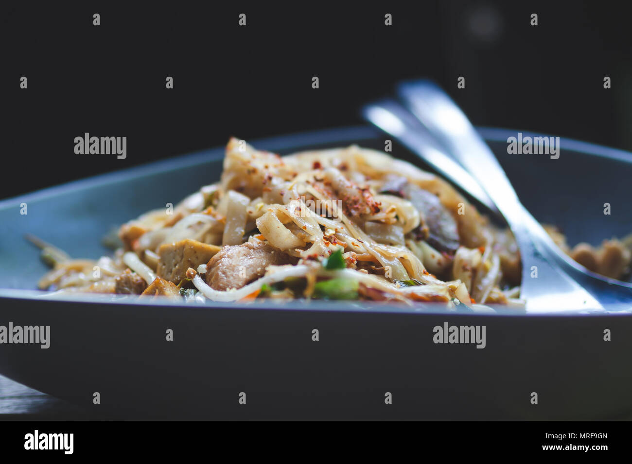 Schüssel Huhn Chow Mein bestreut mit Red Pepper Flakes in einem dunklen Blau Schüssel mit Besteck Stockfoto