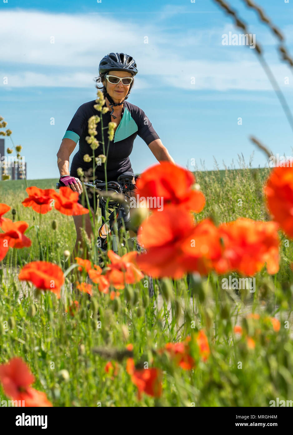 Schöne ältere Frau Reiten e-Mountainbike in einem Vorort einer großen Stadt, umgeben von grünen Feldern und Roter Mohn umgeben Stockfoto