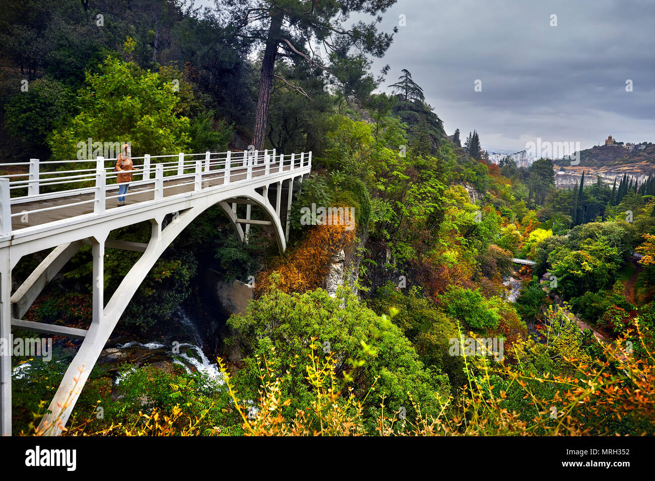 Touristische Frau in Hat an der weißen Brücke im Botanischen Garten mit Bäume im Herbst an bedeckt bewölkten Himmel in Tiflis, Georgien Stockfoto