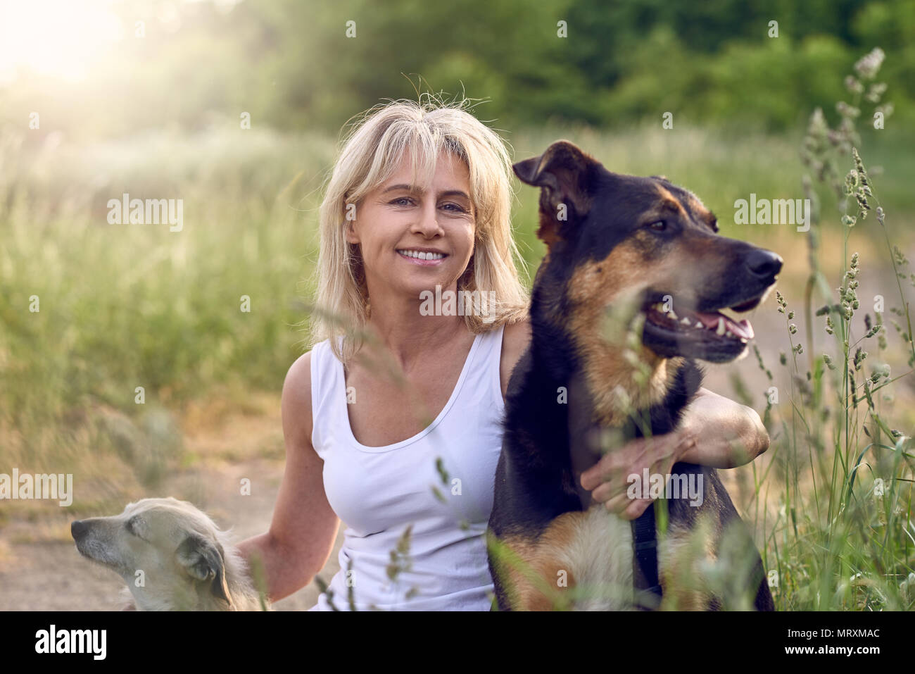 Hübsche blonde Frau mit ihren beiden Hunden hockend mit einem Arm um jedes an der Seite einer ländlichen Straße mit langen Frühling Gras und kopieren Raum auf der rechten Seite Stockfoto