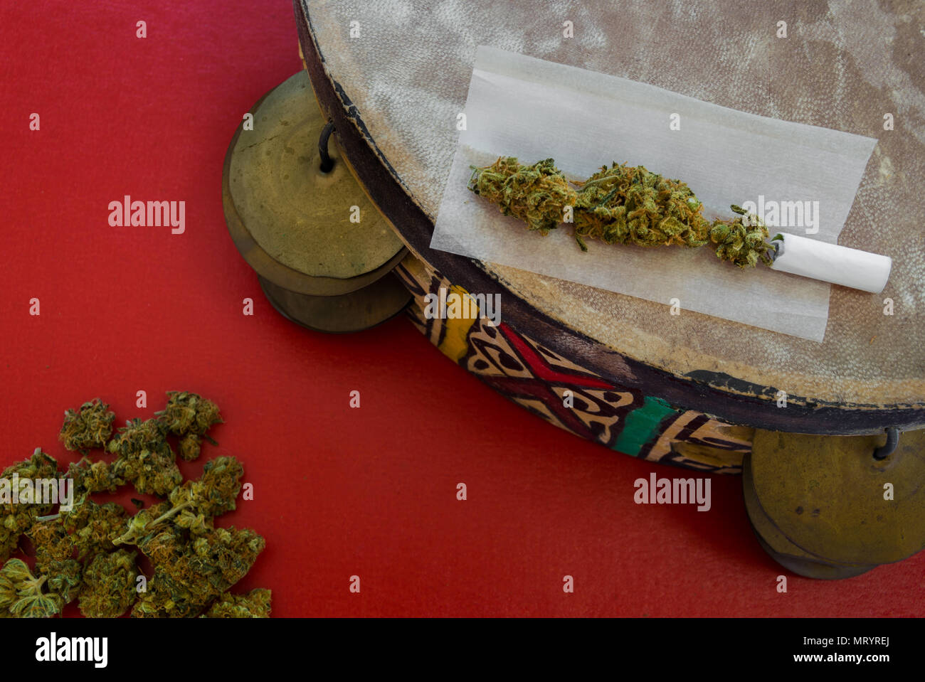 Bunte handgemachte Tamburin mit ethnischen Motiv und eine gemeinsame. Roter Hintergrund mit einem Stapel von Marihuana Knospen. Konzept der Kunst, Drogen und Inspiration. Stockfoto