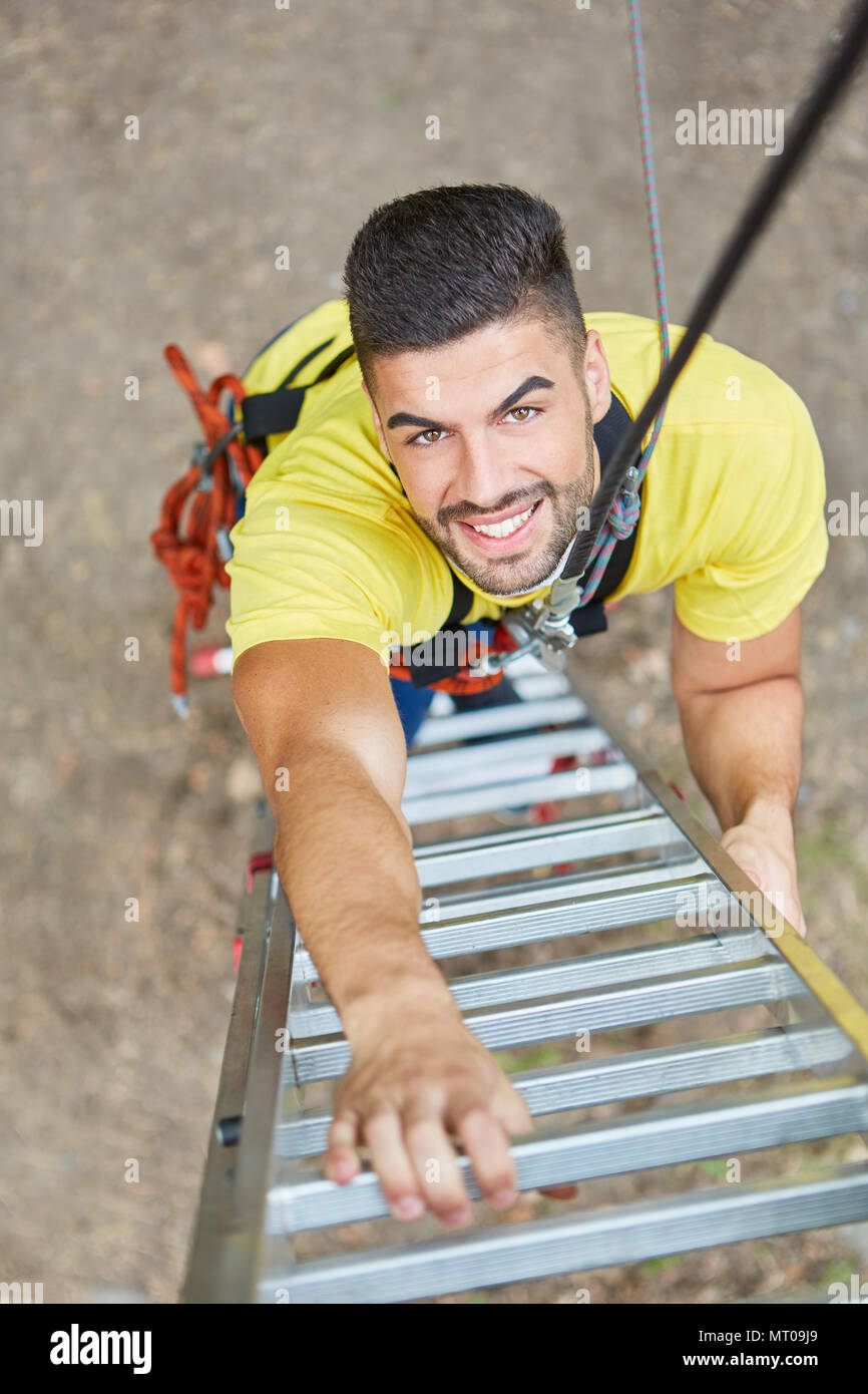 Lächelnden jungen Mann Klettert eine Leiter hinauf in den Hochseilgarten Stockfoto