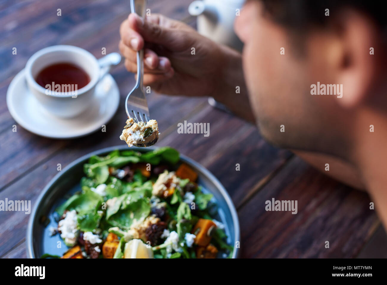 Junger Mann allein sitzen an einem bistrotisch Essen einen leckeren Teller mit gemischter Salat mit einer Gabel und einer Tasse Kaffee Stockfoto