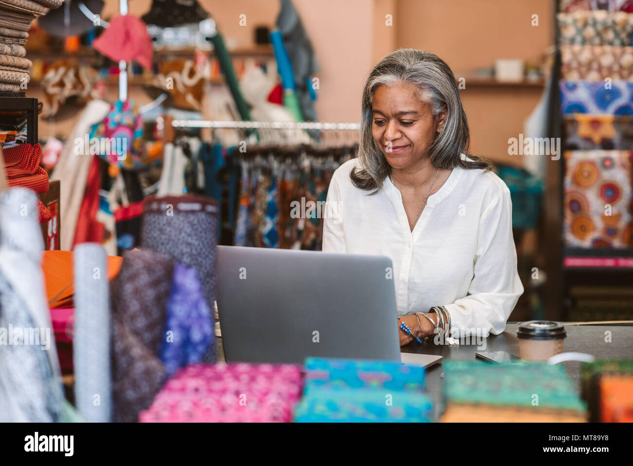 Lächelnd reife Stoff shop besitzer hinter einem Zähler von bunten Tüchern und Textilien umgeben und an einem Notebook arbeitet Stockfoto
