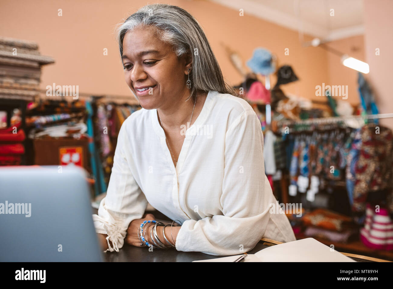 Lächelnd reife Fabric store Inhaber online arbeiten mit einem Laptop an ihrer Ladentheke umgeben von bunten Textilien Stockfoto