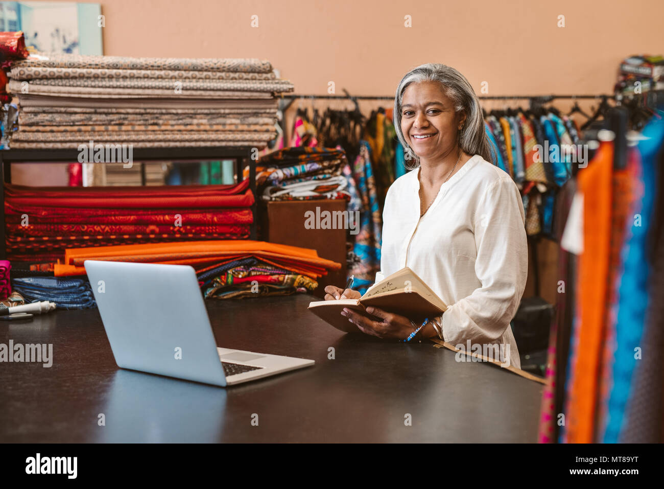 Porträt eines lächelnden Reife fabric Store owner, der an einem Notebook arbeitet und das Schreiben von Notizen bei einem Zähler von bunten Textilien umgeben Stockfoto
