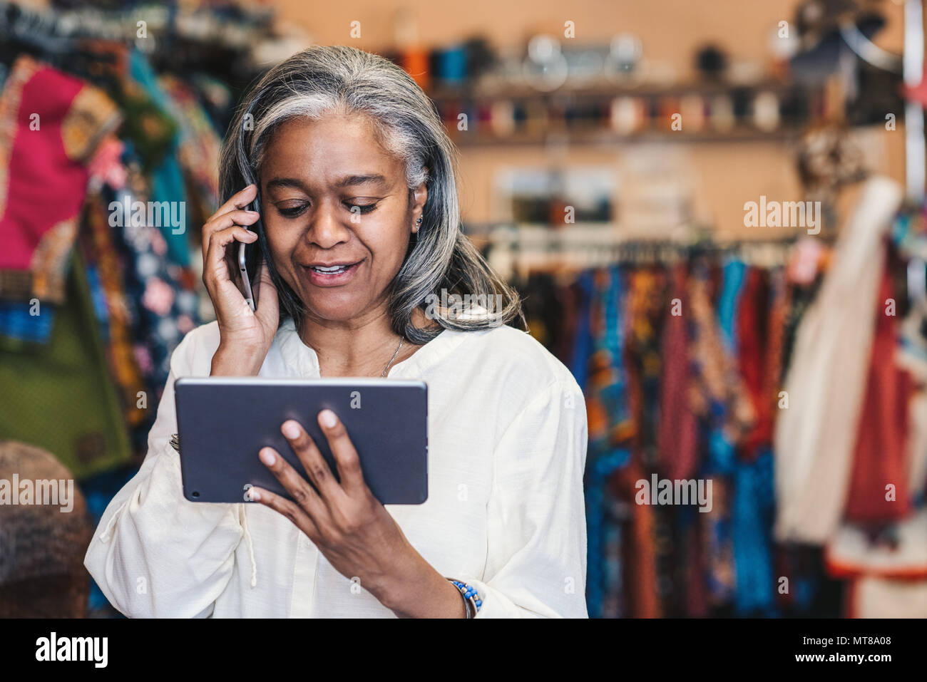 Lächelnd reife Stoff shop besitzer sprechen über ein Handy und die Arbeit mit einem digitalen Tablet stehend von bunten Tüchern und Textilien Stockfoto