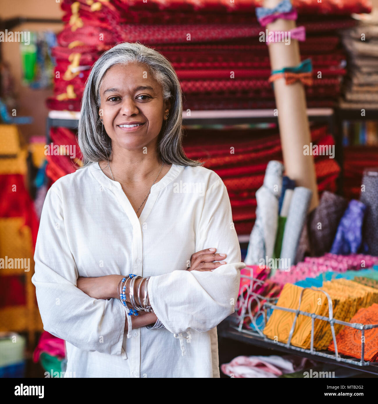 Porträt eines lächelnden reife Gewebespeicher Eigentümer stehen mit ihren Armen von Racks und Regale voll von bunten Tüchern und Textilien gekreuzt Stockfoto