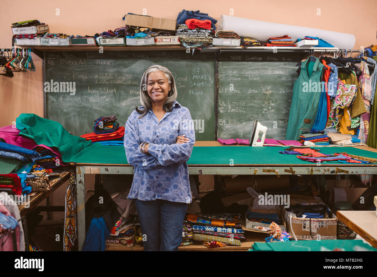 Porträt eines lächelnden reife Näherin stehen mit ihren Armen durch einen Tisch in Ihrem Workshop von bunten Material umgeben gekreuzt Stockfoto