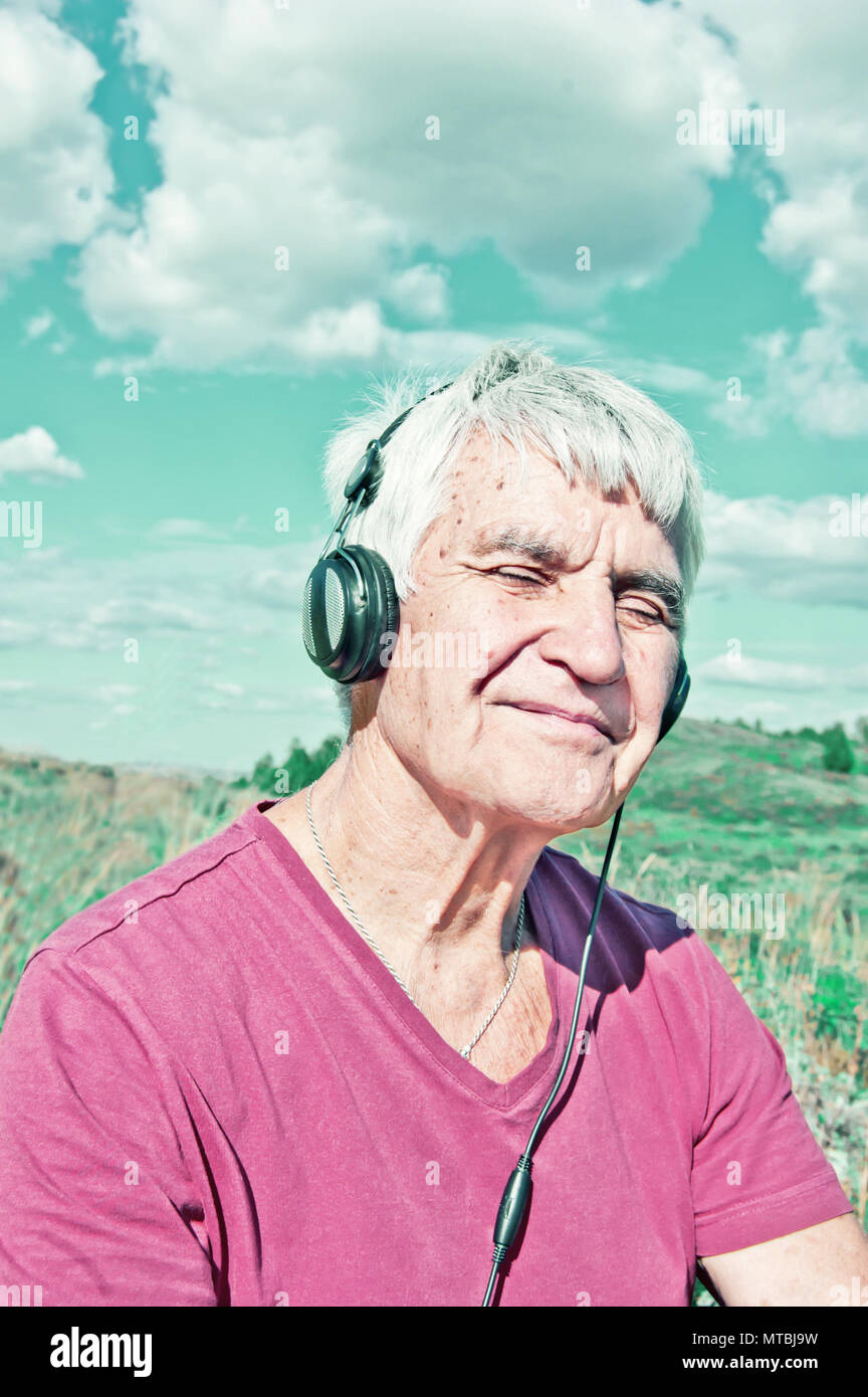 Ein älterer Mann mit geschlossenen Augen über Kopfhörer Musik hört getönten Foto. Ältere lächelnden Mann sitzt im Freien bei bewölktem Himmel. Der Einfluss von Musik auf das Gehirn Stockfoto