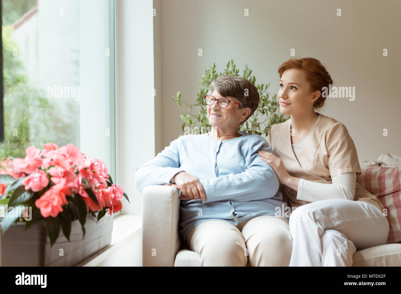 Geriatrische Frau und Ihre professionelle Hausmeister sitzen auf der Couch und schauen durch ein Fenster in einem Wohnzimmer. Blühende Blumen auf der Fensterbank. Stockfoto