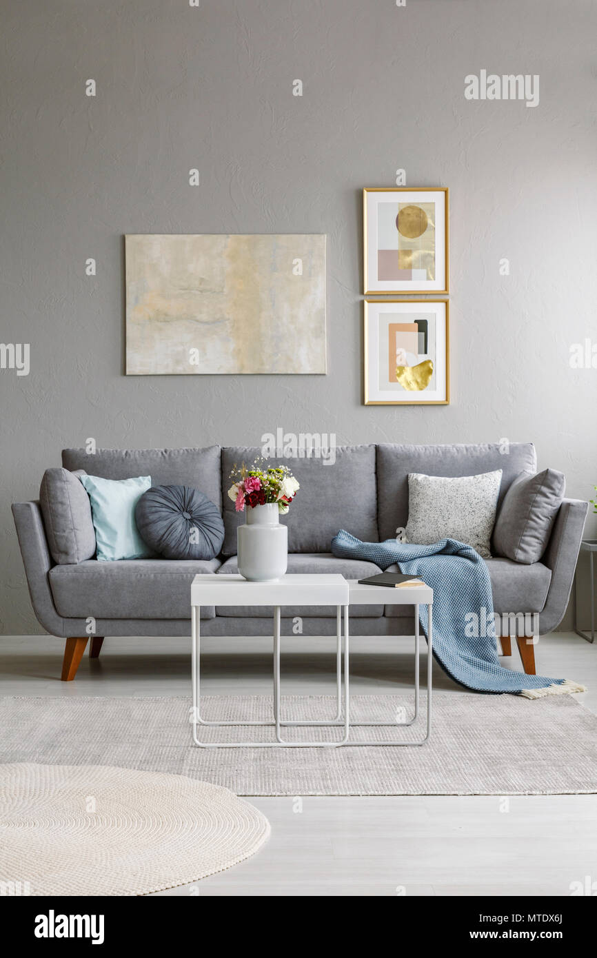 Real Photo von einem grauen Couch mit Kissen und Decke im Wohnzimmer Einrichtung vor einer Wand mit Gemälden und hinter einem weißen Tisch Stockfoto