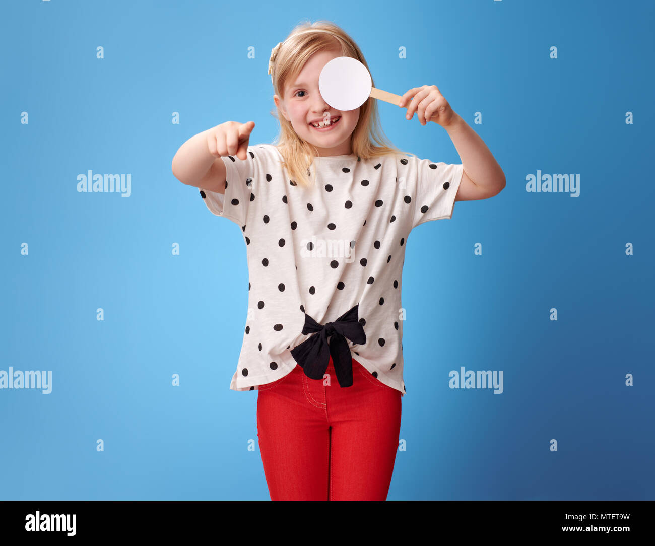 Lächelnde Mädchen in roten Hosen mit einem Auge die Sehschärfe testen und zeigen sie in die Kamera auf blauem Hintergrund isoliert Stockfoto