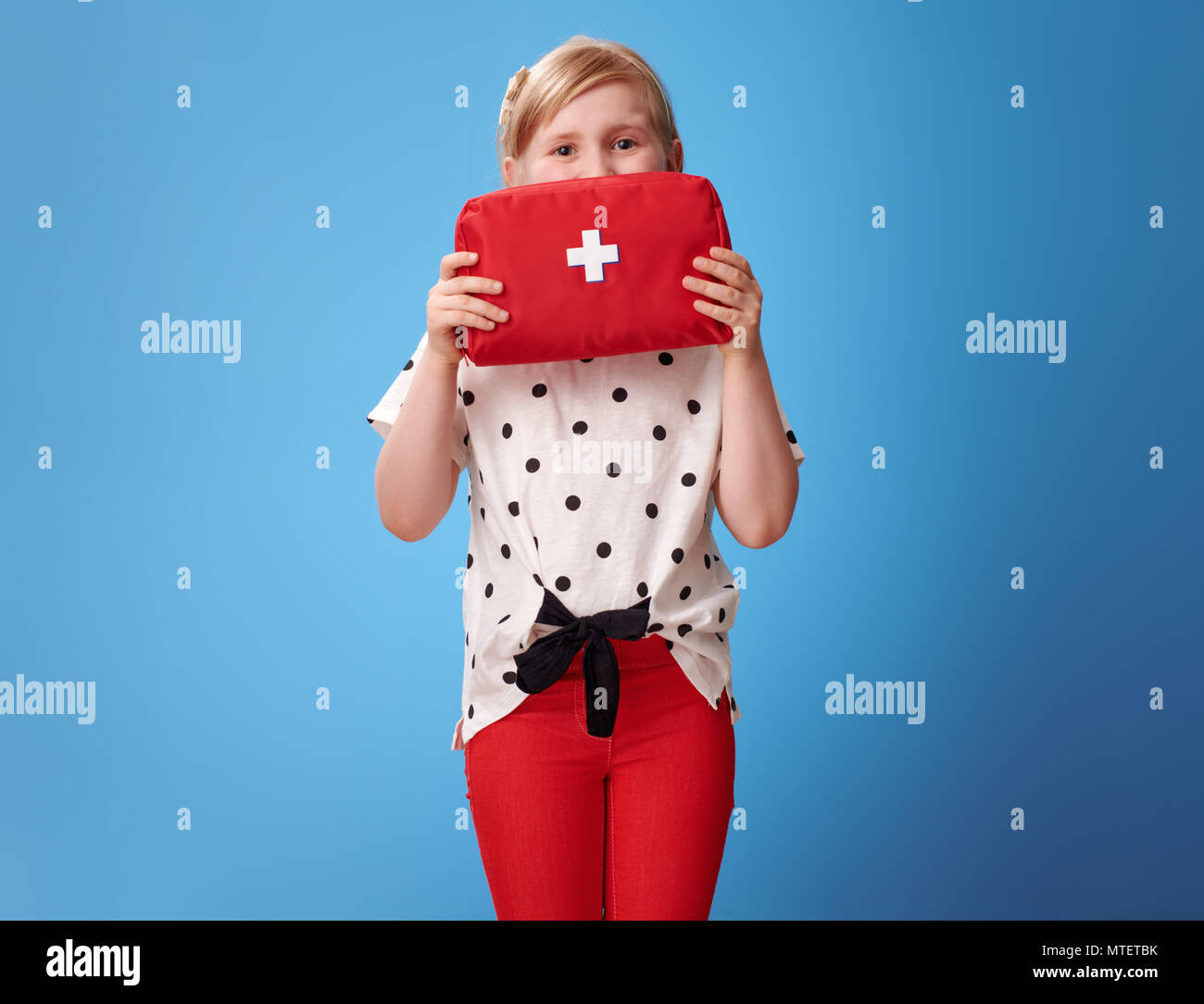 Gerne moderne Kind in roten Hosen holding Erste-Hilfe-Kit vor Gesicht isoliert auf Blau Stockfoto