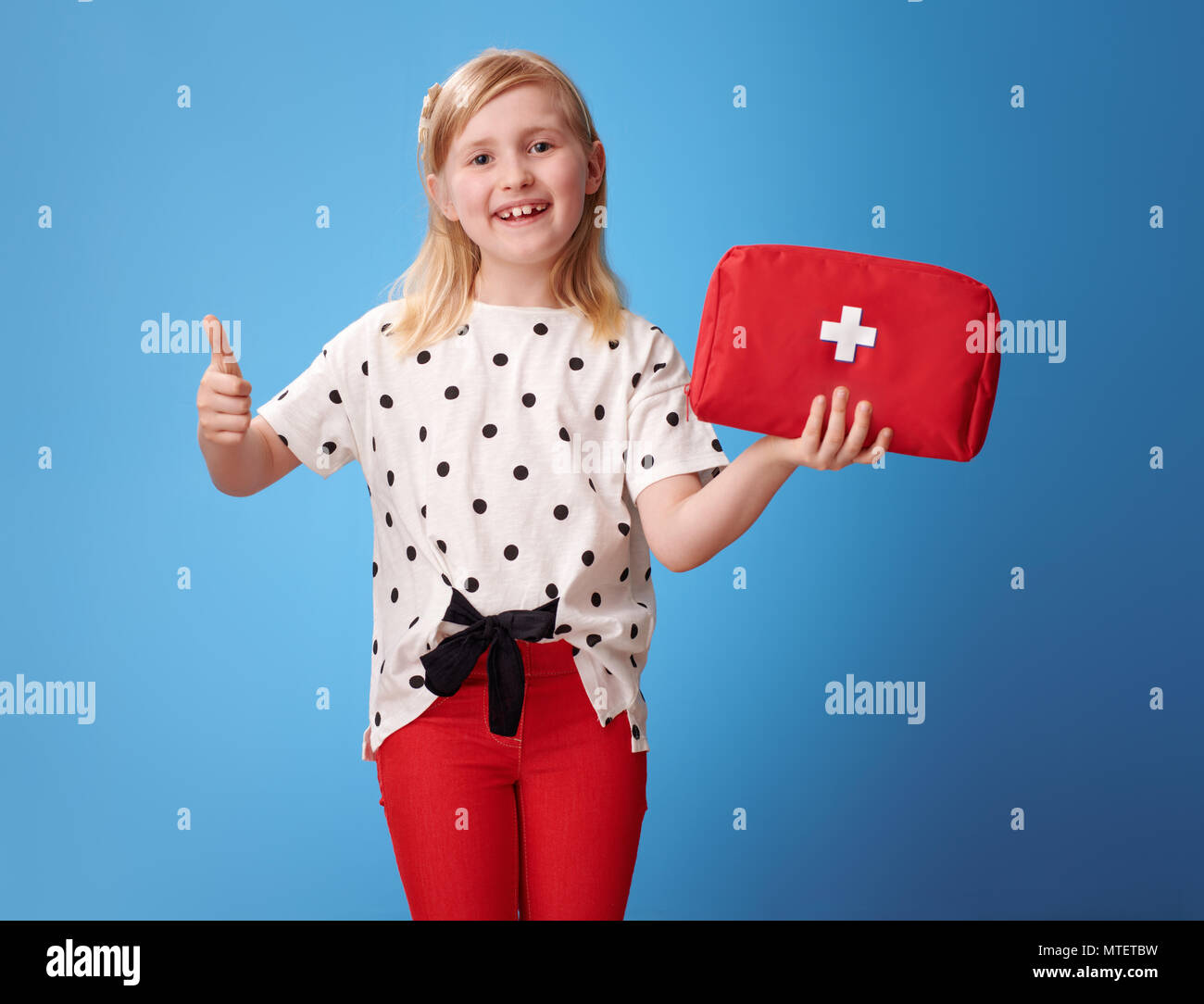 Lächelnd moderne Kind in roten Hosen, Erste-Hilfe-Set und Daumen oben auf Blau isoliert Stockfoto