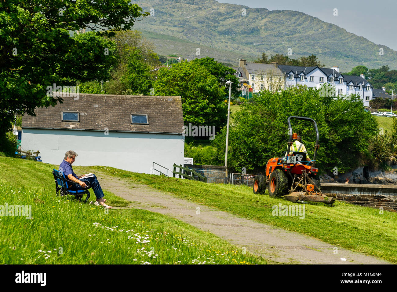County Council Mitarbeiter schneidet das Gras während ein Mann auf einer Bank an einem sonnigen Tag in Schull, County Cork, Irland liest. Stockfoto
