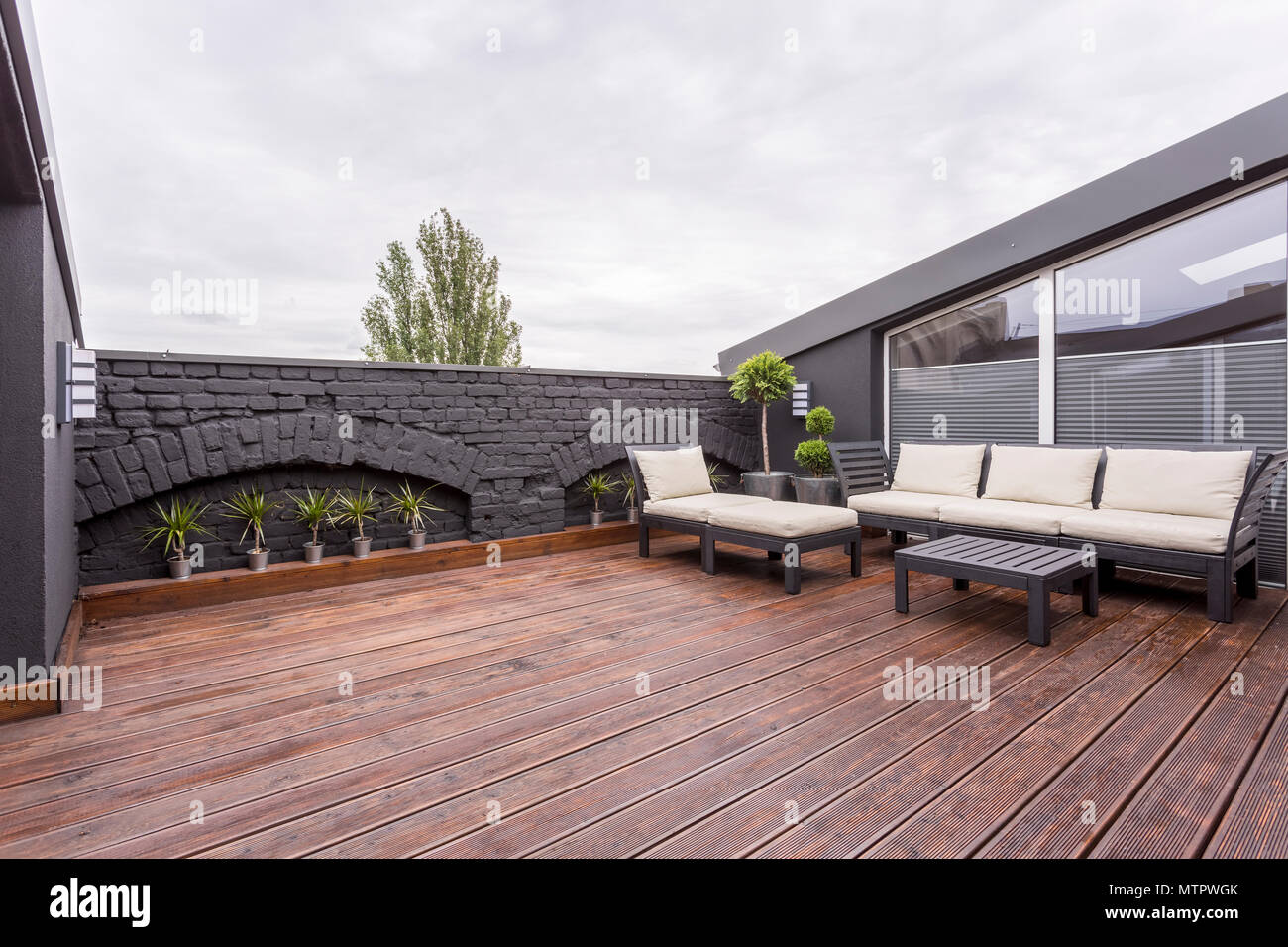 Pflanzen und beige Gartenmöbel auf der Terrasse mit Holzboden und schwarze Wand Stockfoto
