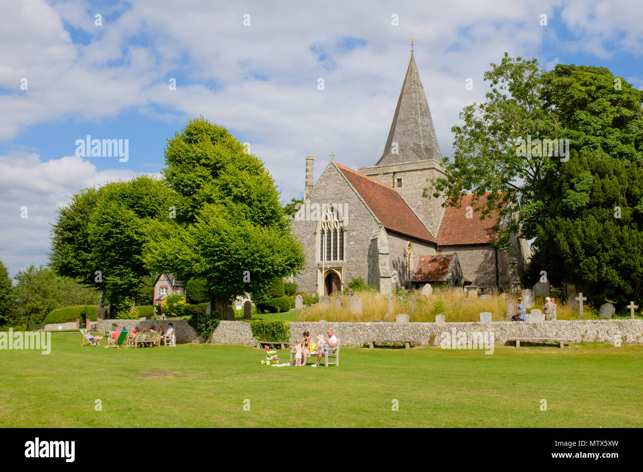 Die Kirche St. Andrew's aus dem 14. Jahrhundert befindet sich auf dem Dorfgrün in Alfriston, East Sussex, England, an einem Sommernachmittag, an dem sich die Menschen auf dem Rasen entspannen. Stockfoto