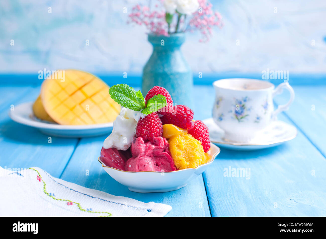 Eis von hellen Farben mit Mango, Himbeeren und Minze in einer Platte, auf blauem Holz- Hintergrund. Eine Tasse Kaffee am Morgen und Licht und Blumen. Vintage Foto. Platz kopieren Stockfoto