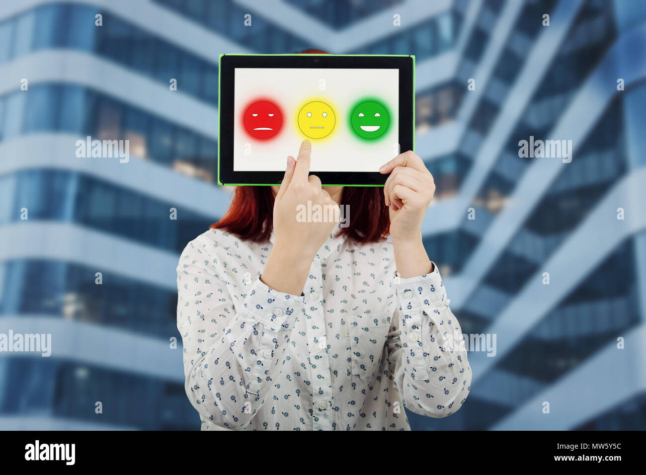 Junge Geschäftsfrau, die ihr Gesicht mit einem digitalen Tablette mit drei Emoticons auf dem Bildschirm, die versuchen, eine Emotion Gesicht zu wählen. Kundenservice ra Stockfoto