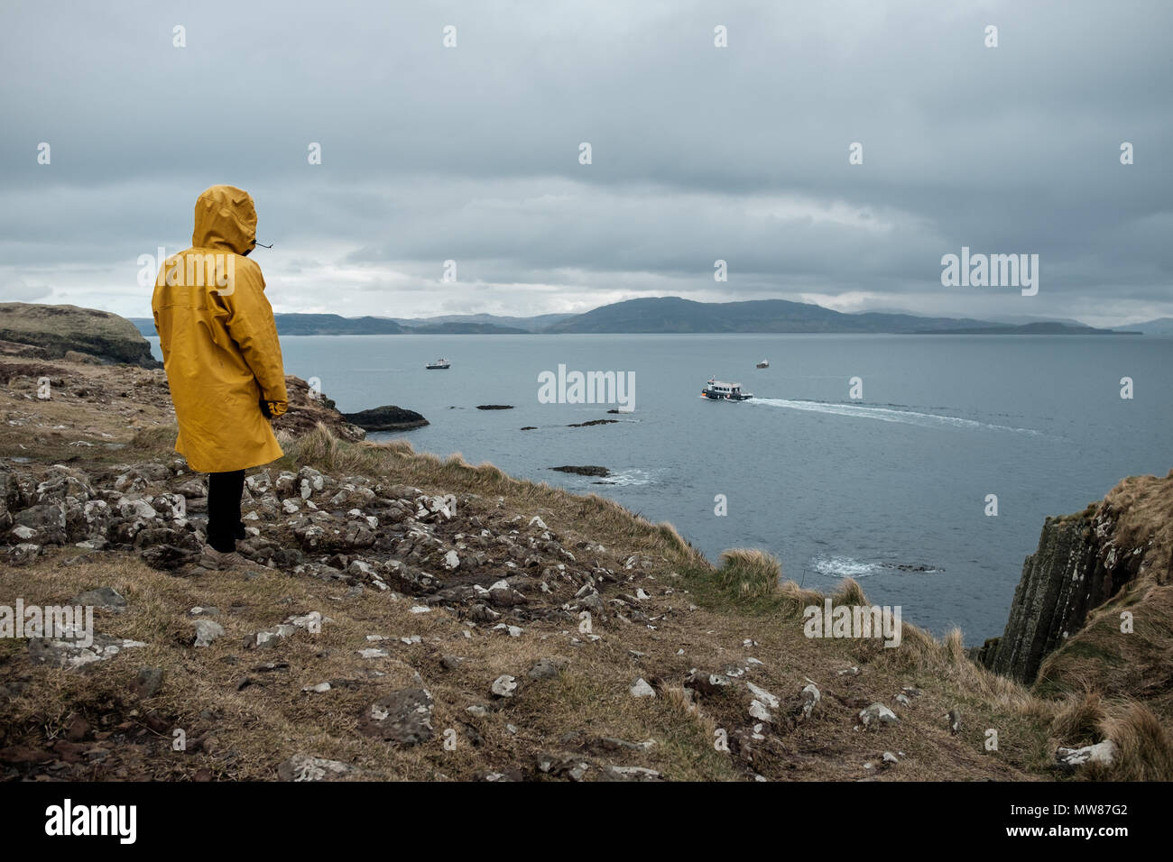 Frau mit Gelb oilskin Jacke beobachten ein Boot von Staffa Insel, in der Nähe der Isle of Mull in Schottland Stockfoto