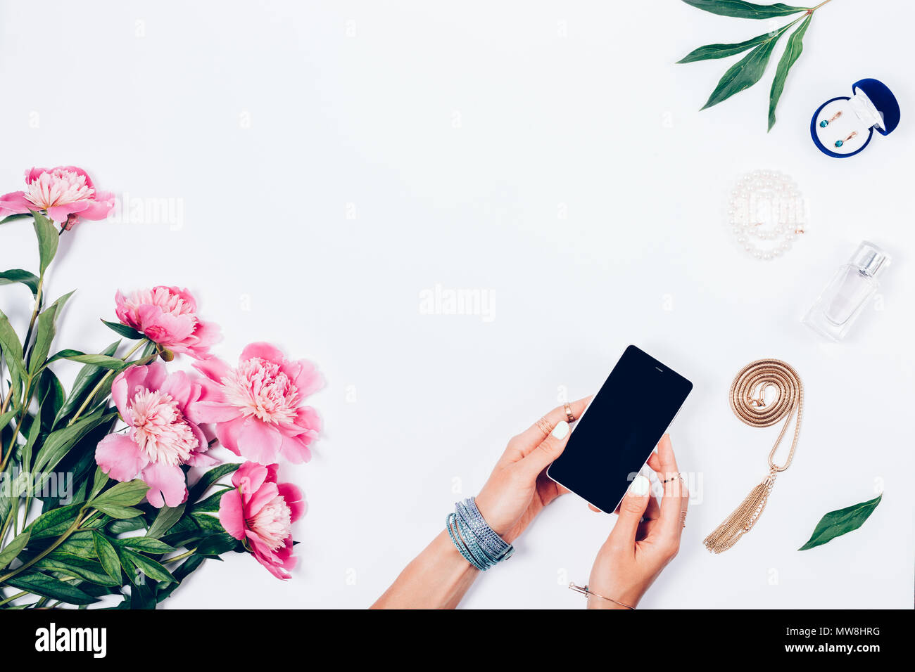 Women's Hände, die Smart Phone auf einem weißen Hintergrund unter Strauß Pfingstrosen und feminin Schmuck. Flach floral Rahmen mit leeren Platz in der c Stockfoto