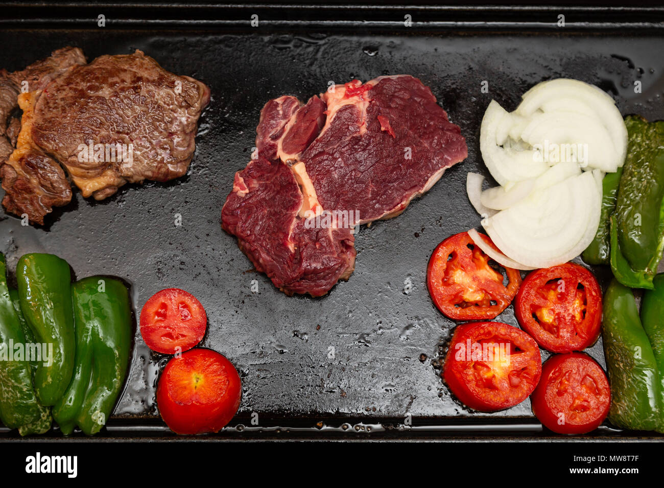 Rib Eye Steak (spanisch: Bife Ancho) roh und gegrillt, Zwiebeln, Tomaten und grüne Paprika auf rechteckiger Gusseisen flache Pfanne, flach legen Stockfoto