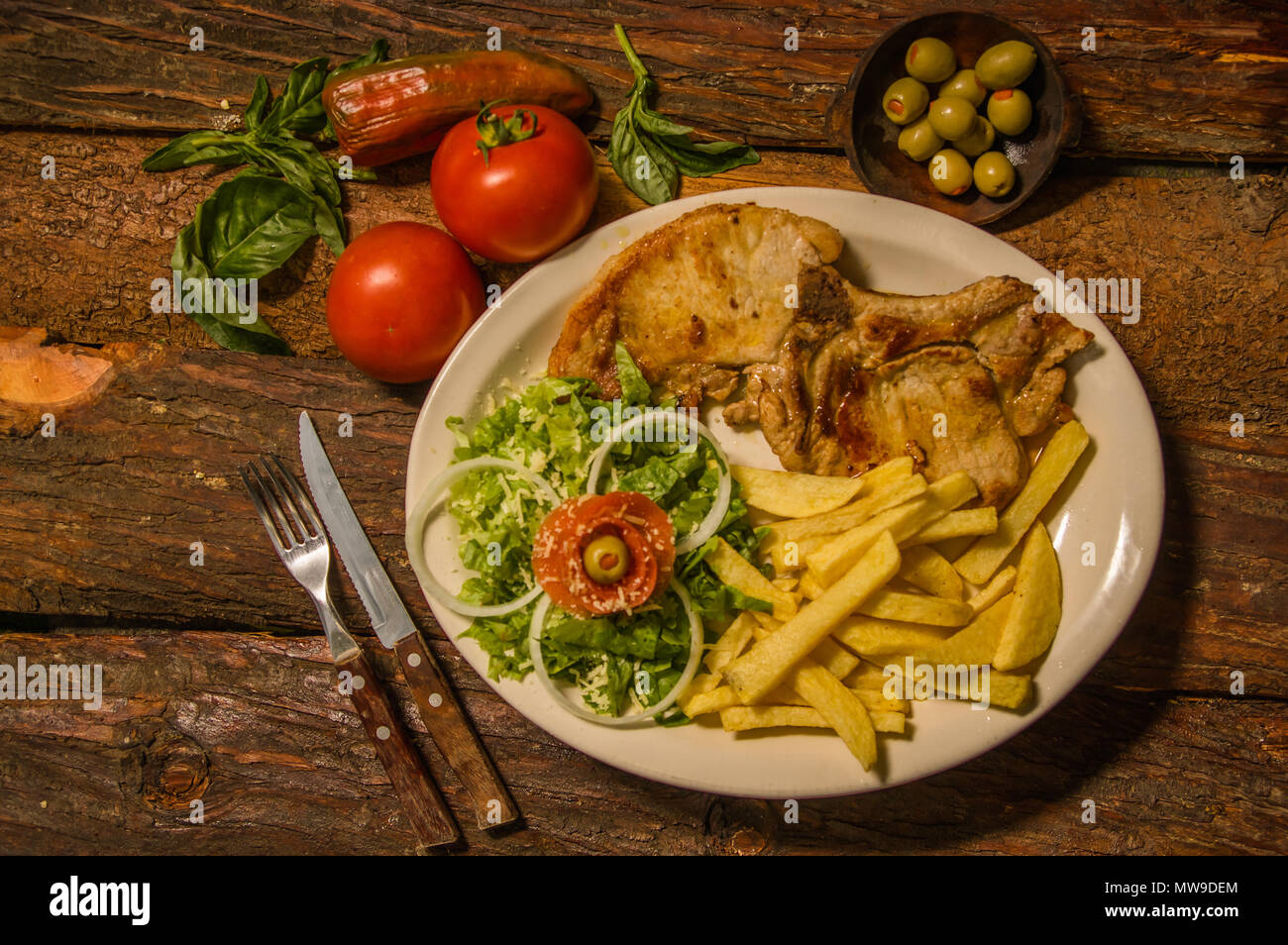 Oben Ansicht von Medium gegrillte Lampe mit Kartoffeln französischen auf weißem Teller serviert, Tomaten, Karotten, Messer und Gabel auf Schneidebrett Chop, auf hölzernen Hintergrund Stockfoto