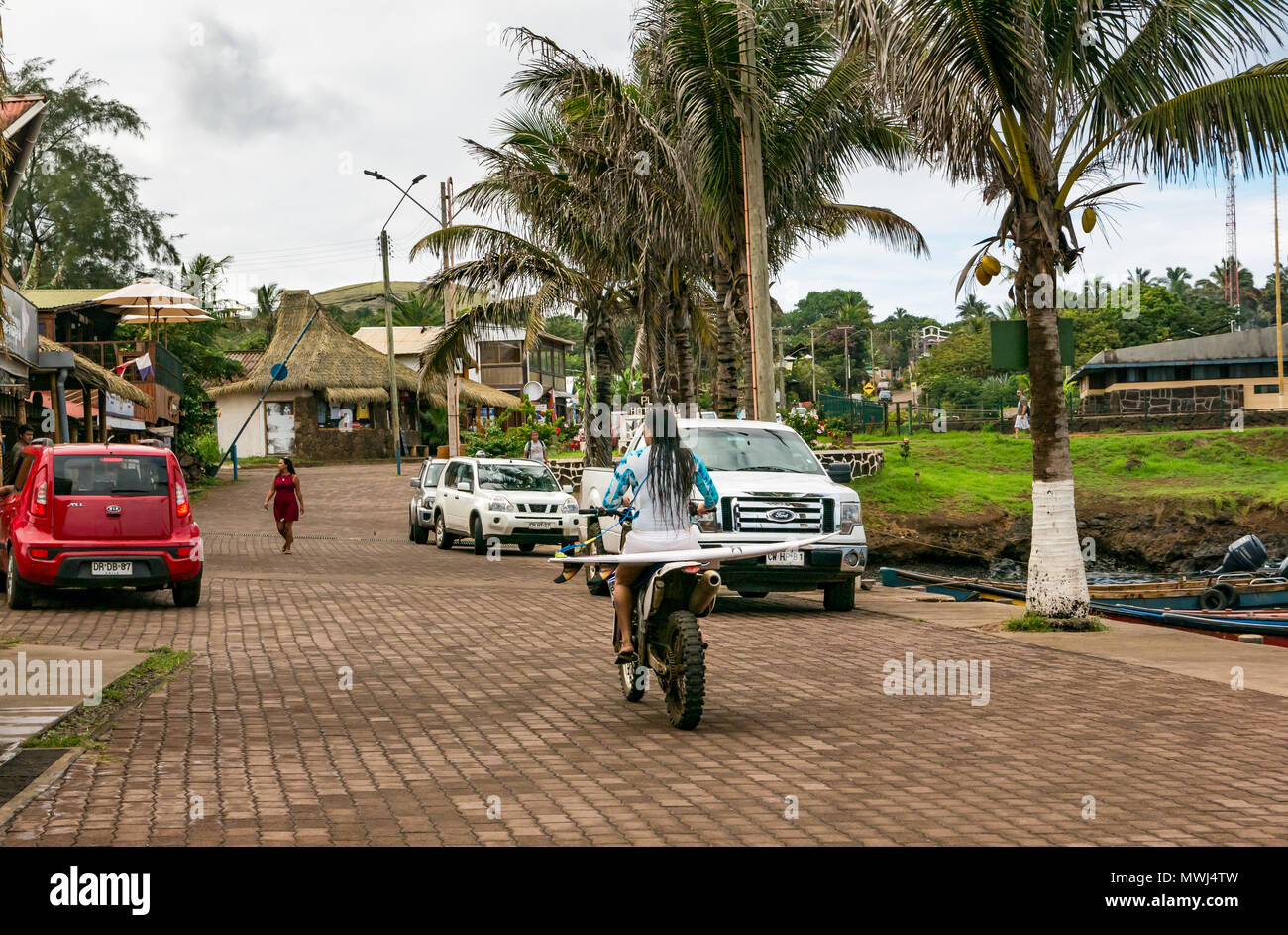 Junge weibliche Surfer tragen Neoprenanzug fahren Motorrad mit Surfbrett auf der Rückseite, Plaza Hotu Matu'a, Hanga Roa, Rapa Nui, Easter Island, Chile Stockfoto