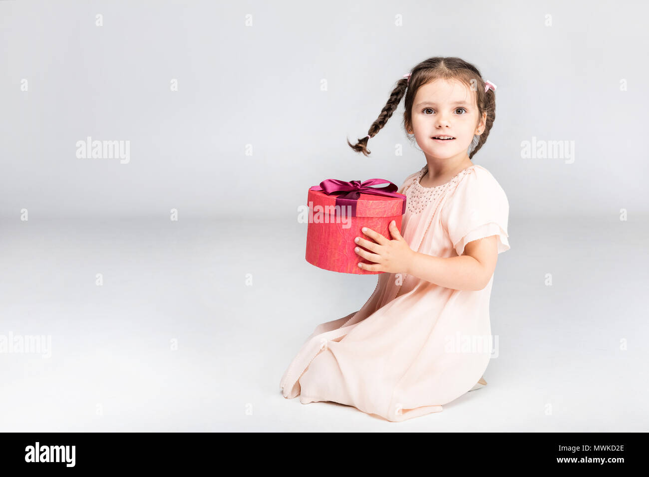 Kleine niedliche Mädchen mit Zöpfen Holding roter Geschenkbox, isoliert auf Grau Stockfoto