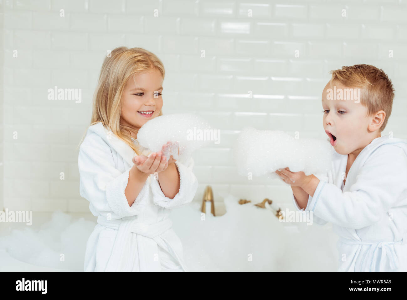 Adorable kleine Kinder in Bademänteln spielen mit Schaumstoff im Badezimmer Stockfoto